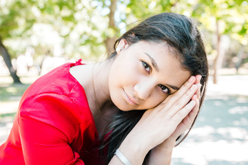 Serene pretty young woman relaxing and posing at camera in park. Beautiful lady wearing red blouse and looking at camera with green trees in background. Relaxation and nature concept. Front view.