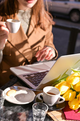 A young girl drinks coffee in a cafe on the street and works on the Internet on a laptop. Sunlight in the frame. Traveling and walking with a noutbuk. Copy space.