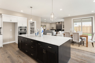 Kitchen and living room interior in new luxury home with open concept floor plan. Features island, hardwood floors, fireplace, dining area, and light filled spaces.