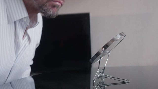 Adult caucasian man sits at table in front of round mirror and examines his eye. MLS