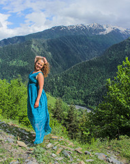 Elegant woman in blue dress at the mountains. Happy girl with wavy hair. Freedom concept. The background of the majestic mountains of the Caucasus and wooden fence. Svaneti, Georgia.
