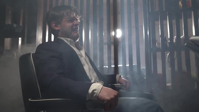 male businessman in blue jacket and white shirt sits in electric chair and twitches. Torture in gloomy place for young guy.