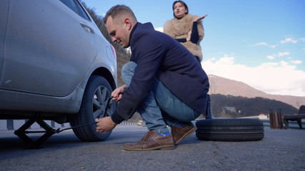 a man changes a wheel on a car, a girl is dissatisfied scolds him