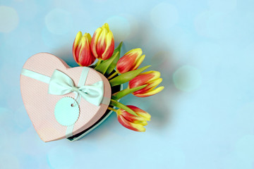 Women day tulips card. Bouquet of yellow tulips in a box in the shape of a heart and a blue bow on a blue background. Copy space, top view, bokeh effect. Congratulatory background for the holiday