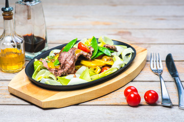 Fried pieces of meat with vegetables in a hot pan on a wooden table in a restaurant.