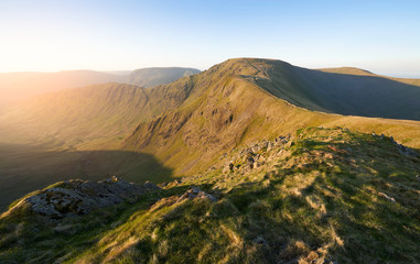 Obraz premium The warm morning glow of sunrise over the summit of High Street with Harter Fell to the left and Thornthwaite to the right from the summit of Rampsgill Head in the Lake District.