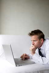 Man in smart casual unbuttoned white shirt lying on white sofa with his laptop computer 