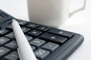 A white pen lies on a black calculator. A white cup is standing nearby. Close-up.