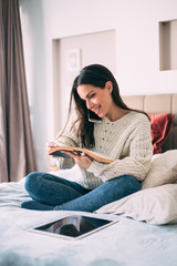 Young beautiful woman caucasian girl reading a book in bed at home student study holding smart phone making call