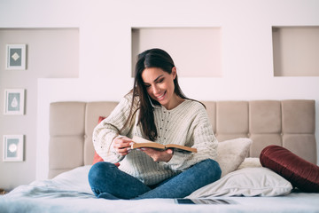 Fototapeta premium Young beautiful woman caucasian girl reading a book in bed at home student study holding smart phone making call