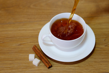 Classic white cup of black tea with saucer, teapot, sugar and cinnamon on the wooden table background. Pouring tea. Breakfast drink at home or cafe.