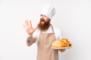 Redhead man in chef uniform. Male baker holding a table with several breads saluting with hand with happy expression