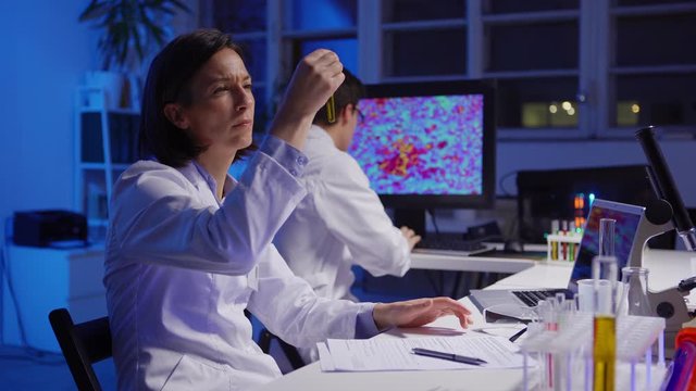 Side view of middle aged female scientist examining liquid in test tube and writing down test results while her male colleague exploring microorganisms on computer screen in laboratory