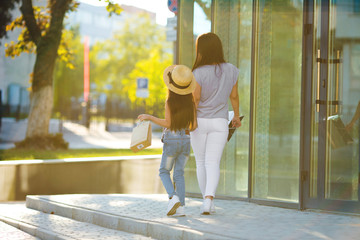 Young mother with cute daughter in hat walking on the street with bags