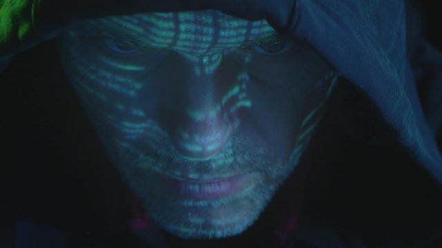 A hacker staring into the computer screen at night with numbers and codes mirroring on his face in dark room with  green light.