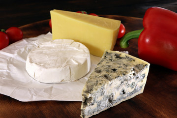 Assortment of different cheese type with tomatoes and sweet pepper on a wooden board. Different pieces of cheese, blue cheese, moldy cheese, camembert and parmesan.
