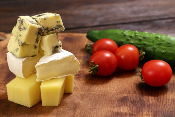 Assortment of different cheese type with tomatoes and cucumber on a wooden board. Different pieces of cheese, blue cheese, moldy cheese, camembert and parmesan.