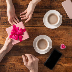 Valentine's Day celebration concept. A nice gift for your loved one. Hands of man and woman with coffee mugs on a wooden table background. Copy space. Flat lay. Close-up.