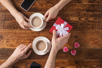Valentine's Day celebration concept. A nice gift for your loved one. Hands of man and woman with coffee mugs on a wooden table background. Copy space. Flat lay. Close-up.