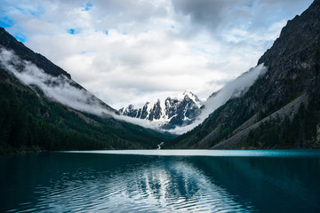 Beautiful big glacier reflected in mountain lake. Rocky snowy mountains, coniferous forest on hills and highland creek under cloudy sky. Atmospheric alpine scenery with low clouds on high steep slopes