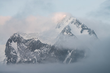 Scenic alpine landscape with snowy mountains inside low clouds at sunrise. Beautiful glacier in dense fog. Soft morning light through clouds. Ghostly scenery with rockies in cloudy sky in pastel tones