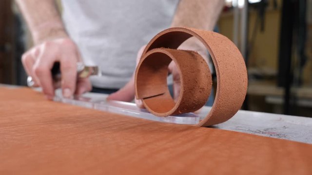 Hands of skilled leather craftsman using plastic pattern and leather knife to cut out part from piece of red leather, close-up shot. Industrial knife man cuts the work piece from the skin. 4 k footage