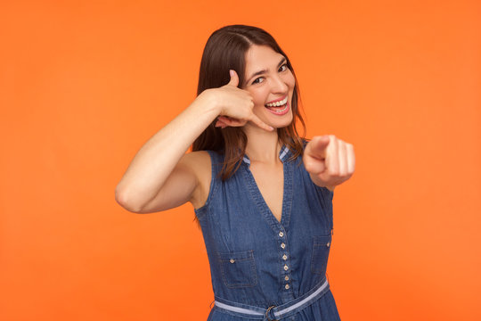Hey you, call me! Happy playful cute brunette woman in denim dress holding arm shaped in telephone gesture and pointing to camera, looking flirtatious. indoor studio shot isolated on orange background