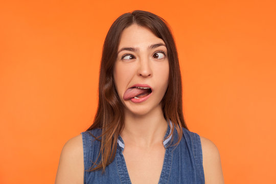 Closeup of silly dumb brunette woman demonstrating tongue and making stupid face with crossed eyes, idiotic expression, showing ridiculous grimace. indoor studio shot isolated on orange background