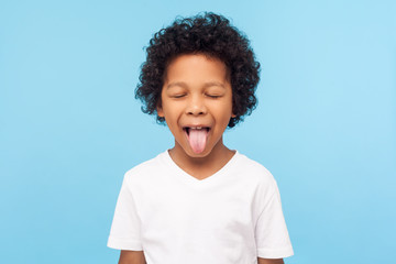 Portrait of funny naughty little boy with curly hair in T-shirt sticking out tongue and keeping...