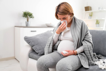 Pregnant woman catching cold, flu, virus, sitting at home on a couch, blowing nose and being sick.