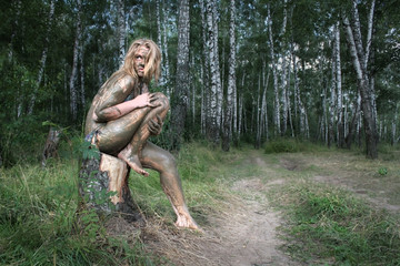A girl with a mud mask sits on a birch stump at an ecological resort. Beautiful blonde - dirty, wild and alone in the forest