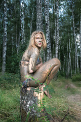 A girl with a mud mask sits on a birch stump at an ecological resort. Beautiful blonde - dirty, wild and alone in the forest