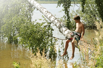 Naklejka na ściany i meble A tanned boy of 12 years old is sitting on a birch trunk that has fallen. Russian