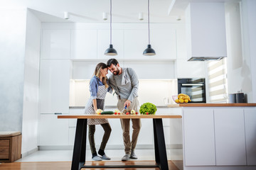 Young attractive caucasian bearded man hugging his beloved girlfriend and cutting vegetables for meal. Healthy eating concept.