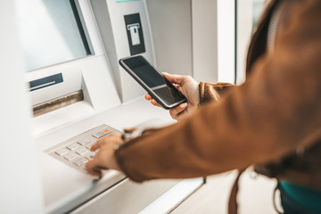 Close up shot of young adult woman. He typing pin code on keypad of ATM machine while using smart...