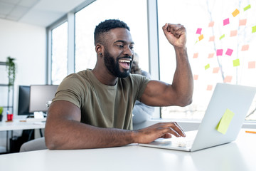 A young man celebrating success he learned about via lap top in an indoor environment.