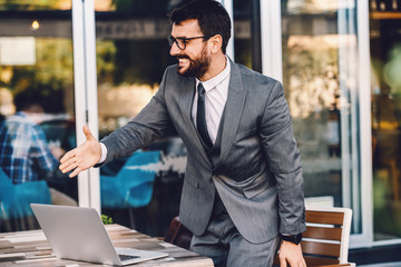 Young smiling cheerful friendly caucasian bearded sophisticated businessman in suit with eyeglasses standing in cafe and extending his hand for handshake with his business partner.