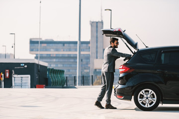 Full length of handsome caucasian bearded rich businessman in suit opening trunk on his car and...