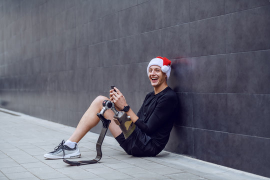 Cheerful Caucasian sportsman with artificial leg and with santa hat on head sitting on ground, leaning on wall and sending wishes for christmas over his smart phone.