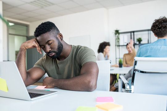 A man slacking at work, too tired. There are a few colleagues behind him in the office.