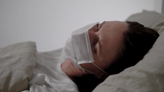 sick woman with medical mask is coughing hardly lying in bed in hospital room, closeup of face