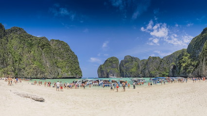 view seaside of many touris walking on white sand beach with many speed boats floating in blue-green sea around with high mountains and blue sky background, Maya Bay, Phi Phi islands, Krabi, Thailand.