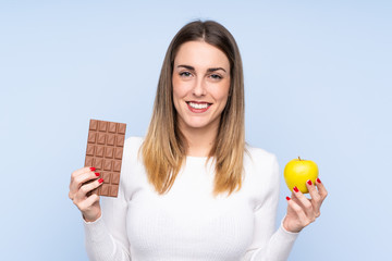 Young blonde woman over isolated blue background taking a chocolate tablet in one hand and an apple in the other