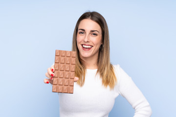 Young blonde woman over isolated blue background taking a chocolate tablet and happy