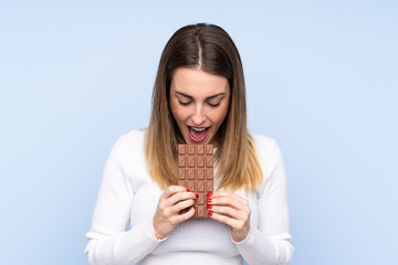 Young blonde woman over isolated blue background eating a chocolate tablet
