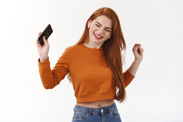 Fototapeta premium Rock-on. Excited and carefree attractive modern redhead woman having fun, enjoying awesome music favorite artist, made playlist, dancing raising hand up with smartphone, wear wireless earphones