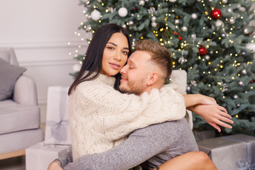 young beautiful couple, brunette girl in a white sweater, a blond man in a gray sweater sitting near a Christmas tree