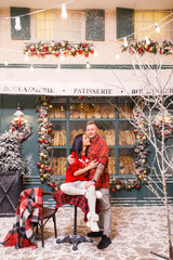 Young beautiful couple, a brunette girl in a red sweater and white pants, a man in a red shirt are sitting on the streets of Paris against the background of a window of a bakery shop