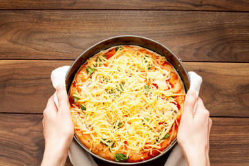 The pizza is completely strewn with cheese, before being sent to the oven in hands in a potholder, on a wooden background. The concept of cooking pizza, homemade recipes for food.
