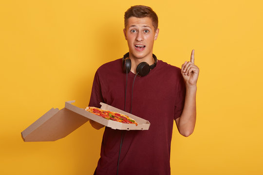 Horizontal shot of pizza delivery man wearing casual clothing holding pizza boxes over isolated yellow background with index finger up, having good idea, handsome guy having astonish fasial expression